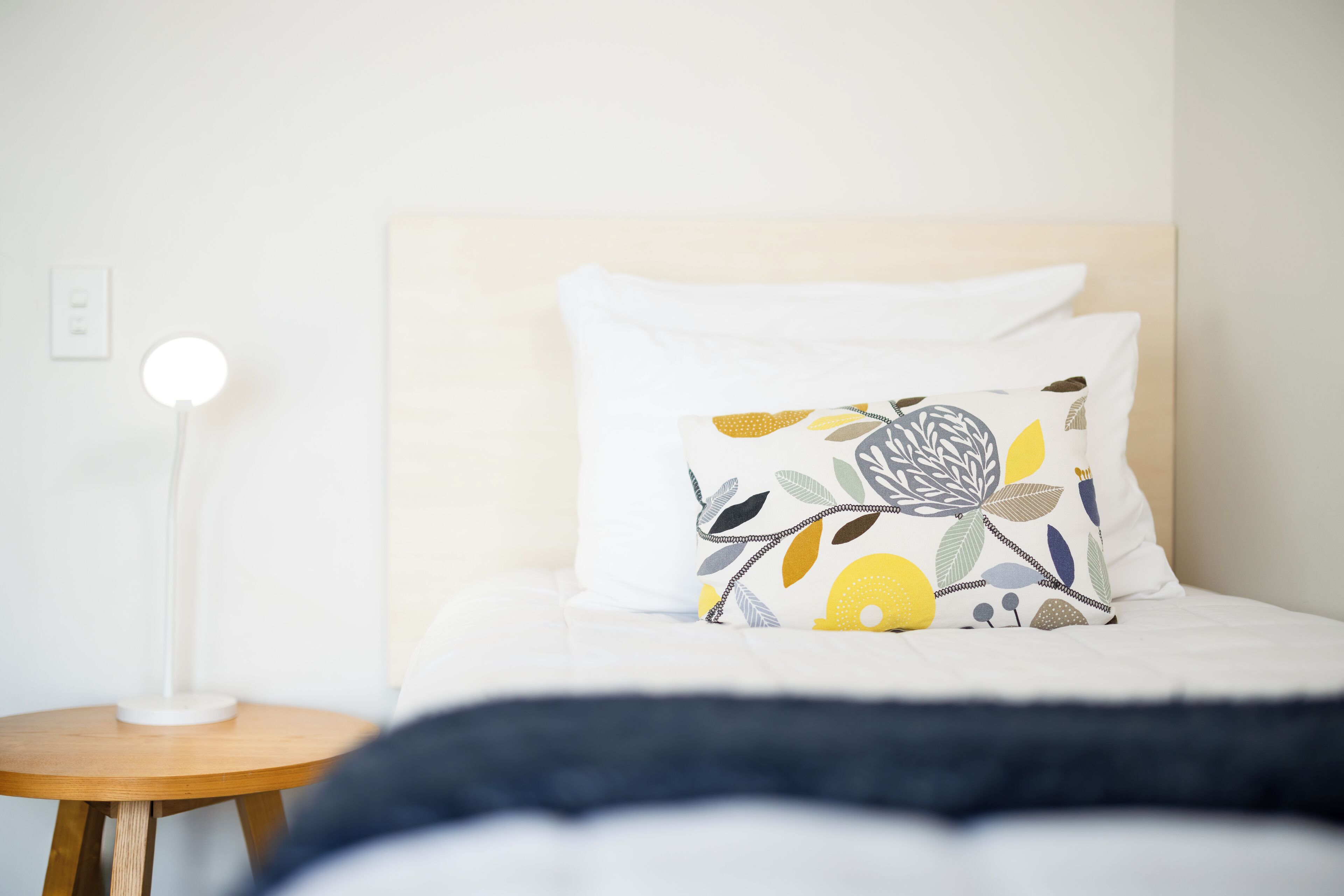 Een netjes opgemaakt bed met een wit kussen en een decoratief kussen met een bladmotief. Er staat een rond tafeltje met een witte lamp naast het bed.
