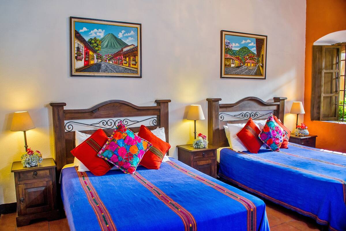 A hotel room with two beds, each with a blue bedspread and red pillows, wooden headboards, and wall-mounted paintings.