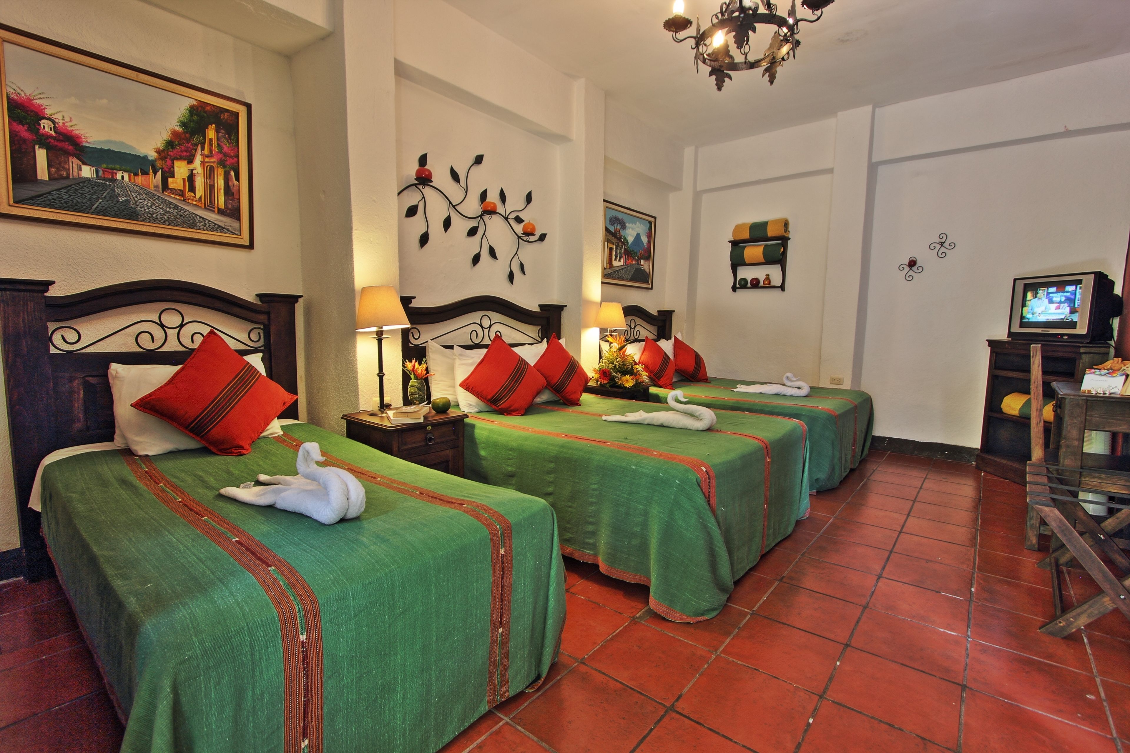 A hotel room with three beds, each with a green bedspread and red pillows, a chandelier, and a television on a wooden stand.
