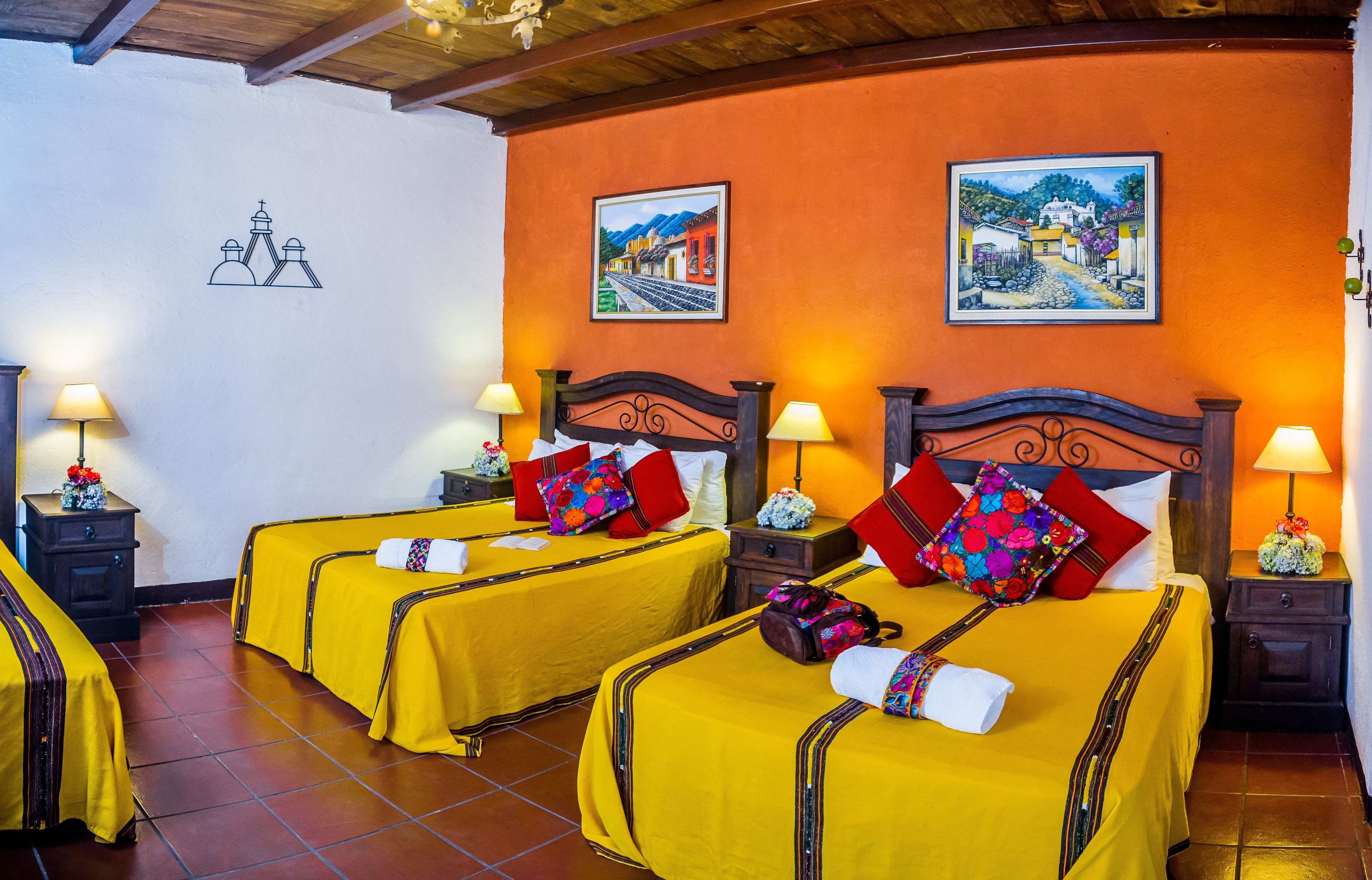 Habitació d'hotel amb dos llits, cadascun amb roba de llit groga i coixins vermells, panells de capçal de fusta i làmpades laterals.