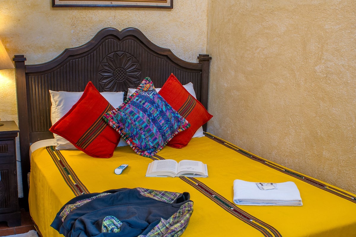 A neatly made bed with a yellow bedspread, red and multicolored pillows, and a book and remote control on it.