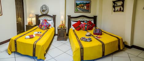 Two beds with yellow bedsheets and decorative pillows, a small table with a lamp, and a framed picture on the wall.