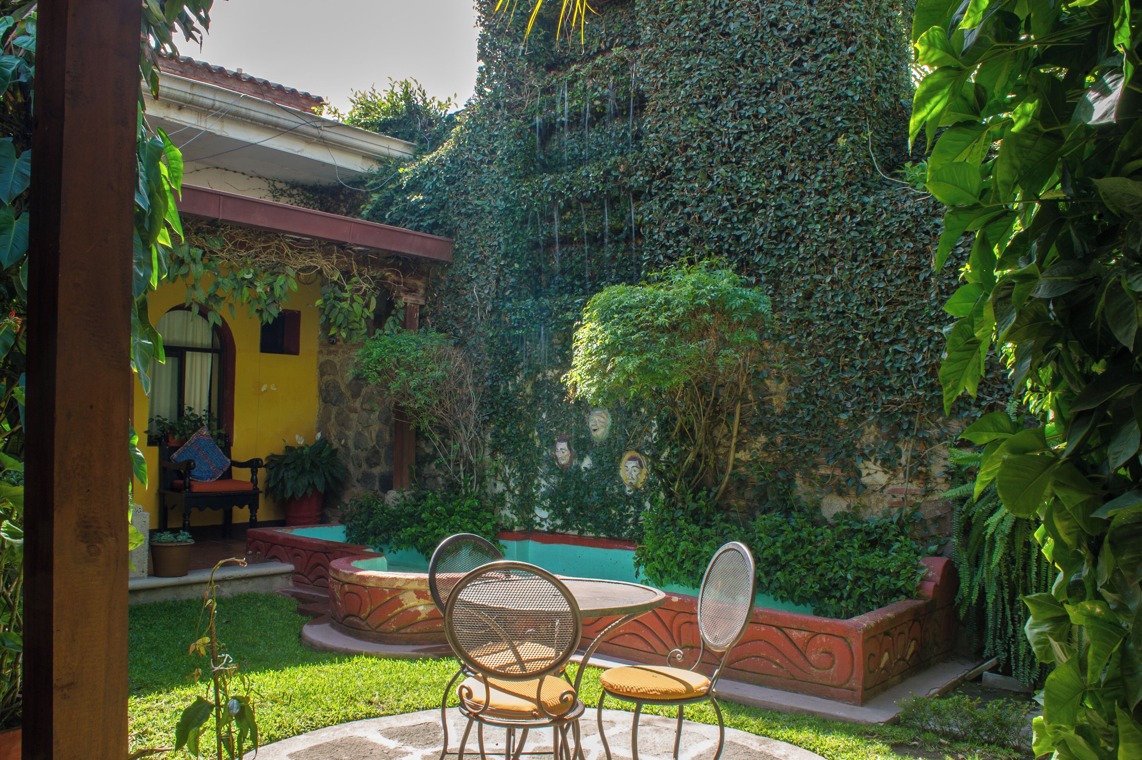 A courtyard with a round table and two chairs, surrounded by lush greenery and a stone wall.