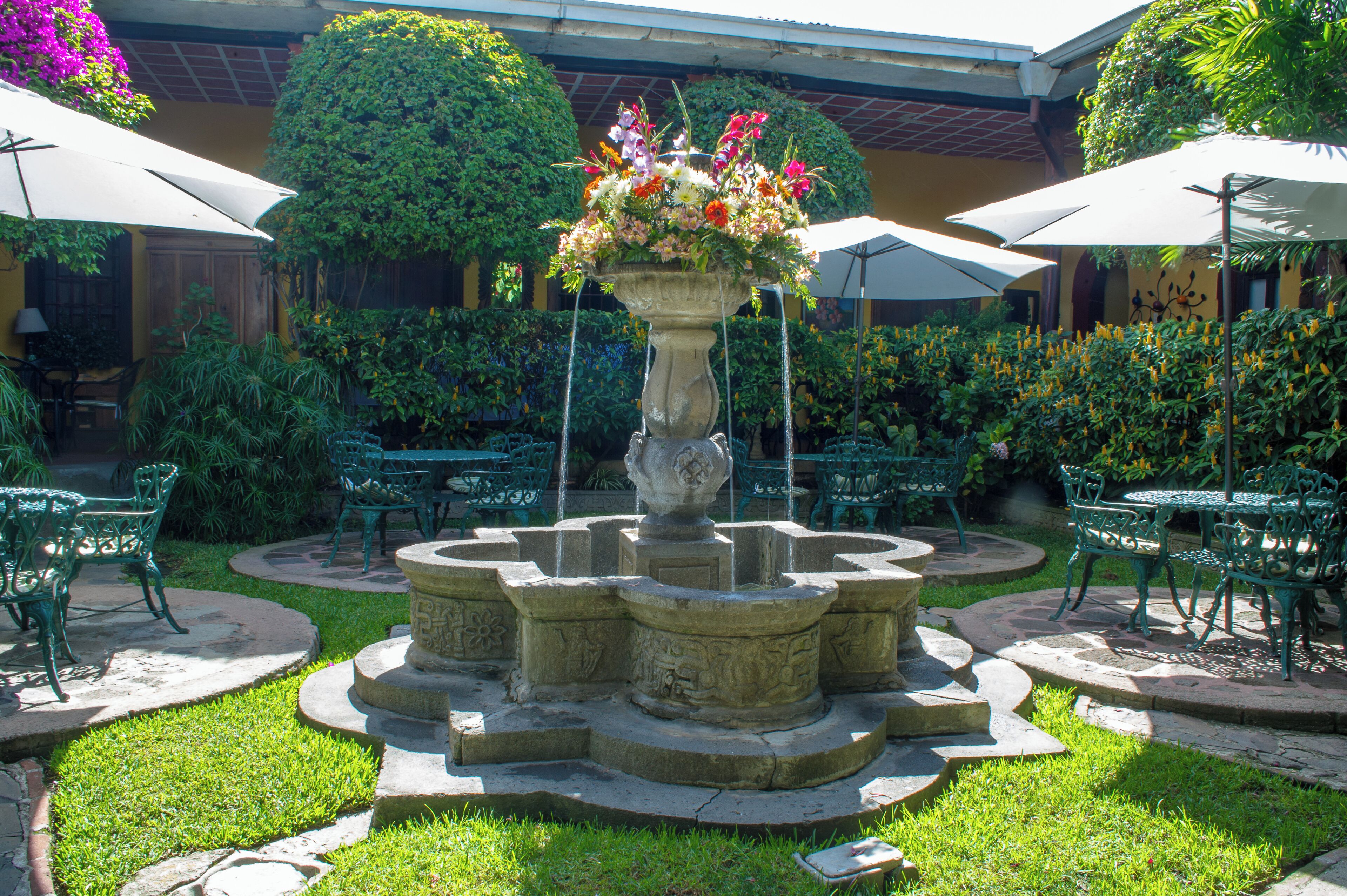 A courtyard with a central fountain, surrounded by green chairs and tables under umbrellas.