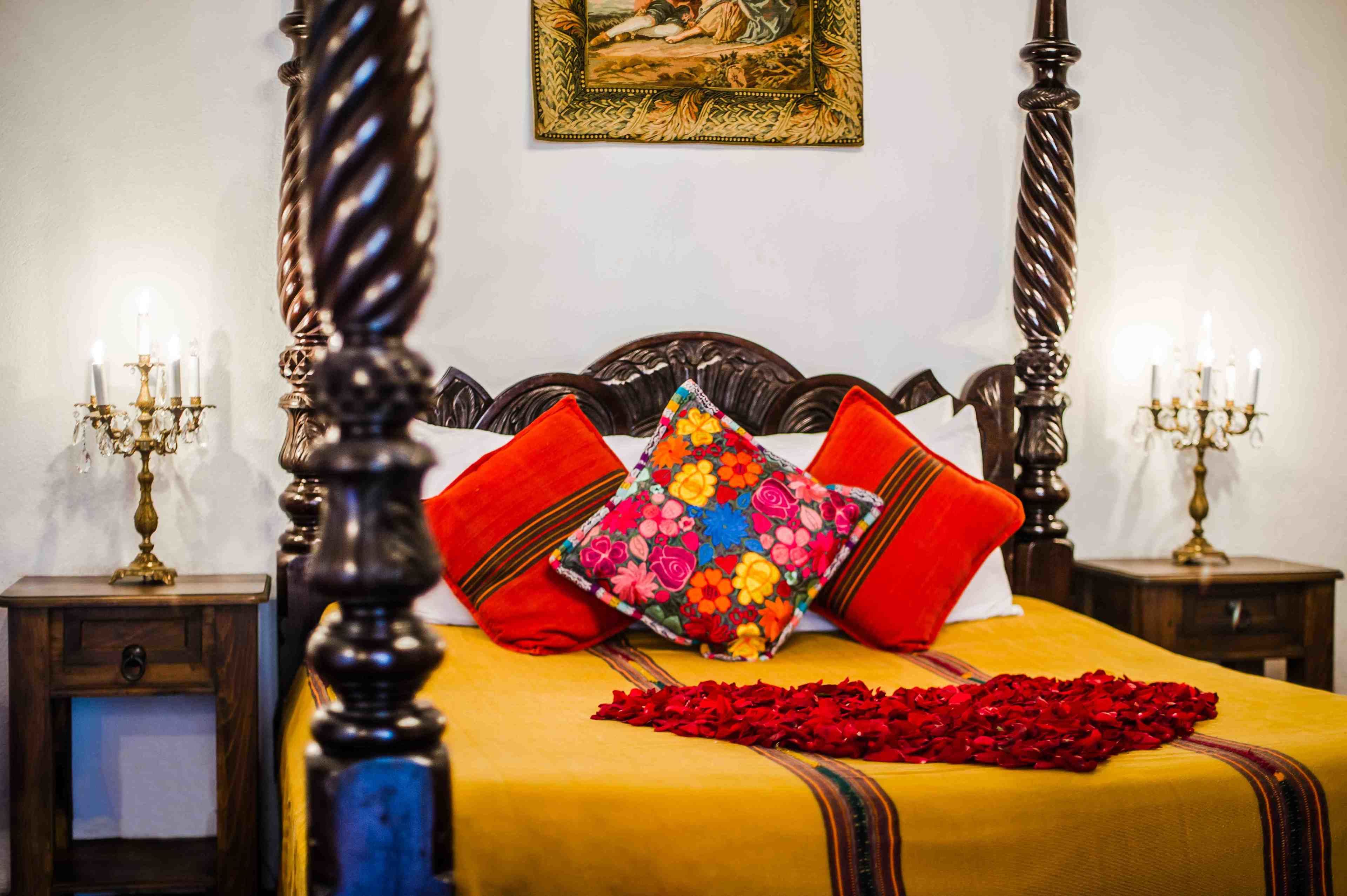 A four-poster bed with a yellow bedspread and red pillows, a red rose petal arrangement, and a framed picture on the wall.