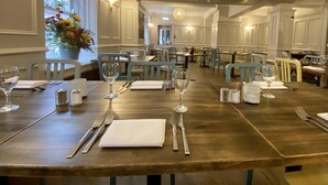 A dining area with wooden tables, white chairs, and neatly set tables with cutlery and wine glasses.