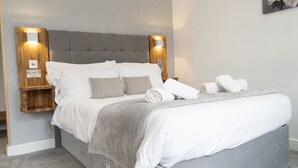A neatly made bed with white and grey linens, a grey upholstered headboard, and two bedside tables with wall-mounted lamps.