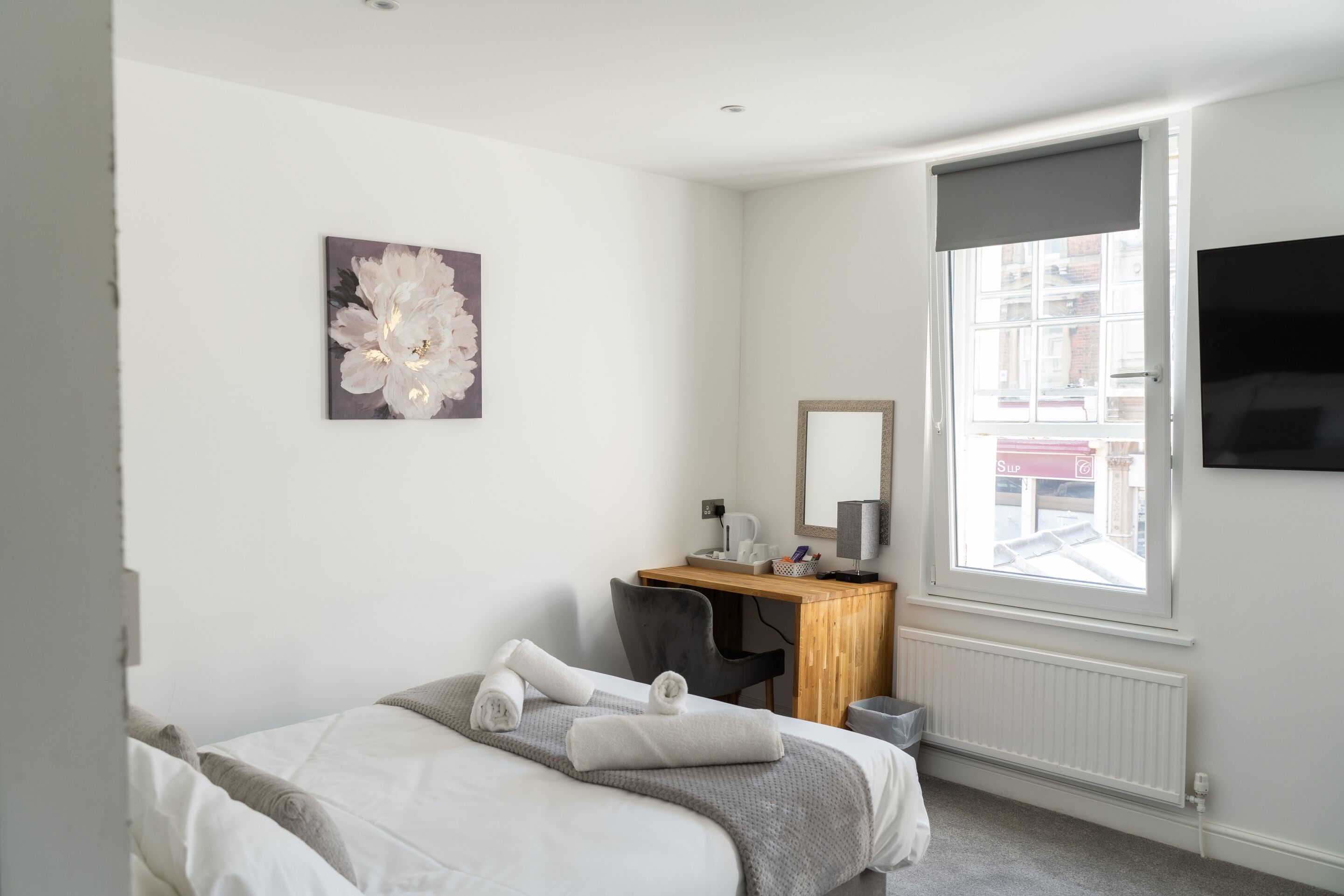 A hotel room with a bed, a desk, a chair, a large window, a flat-screen TV, and a wall-mounted artwork.