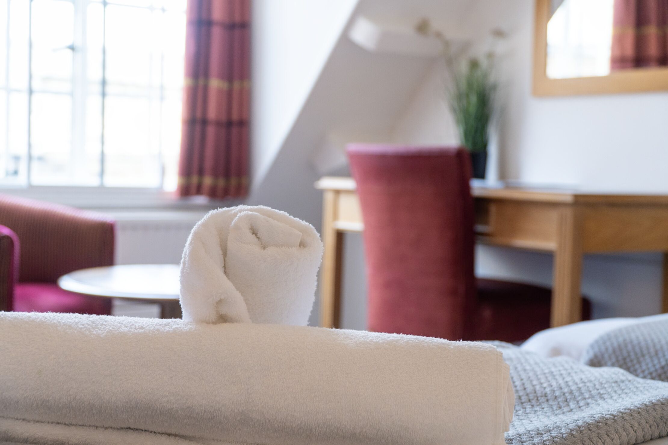 Una cama bien hecha con una toalla blanca, una silla roja y una pequeña mesa redonda en una habitación con ventana y planta.