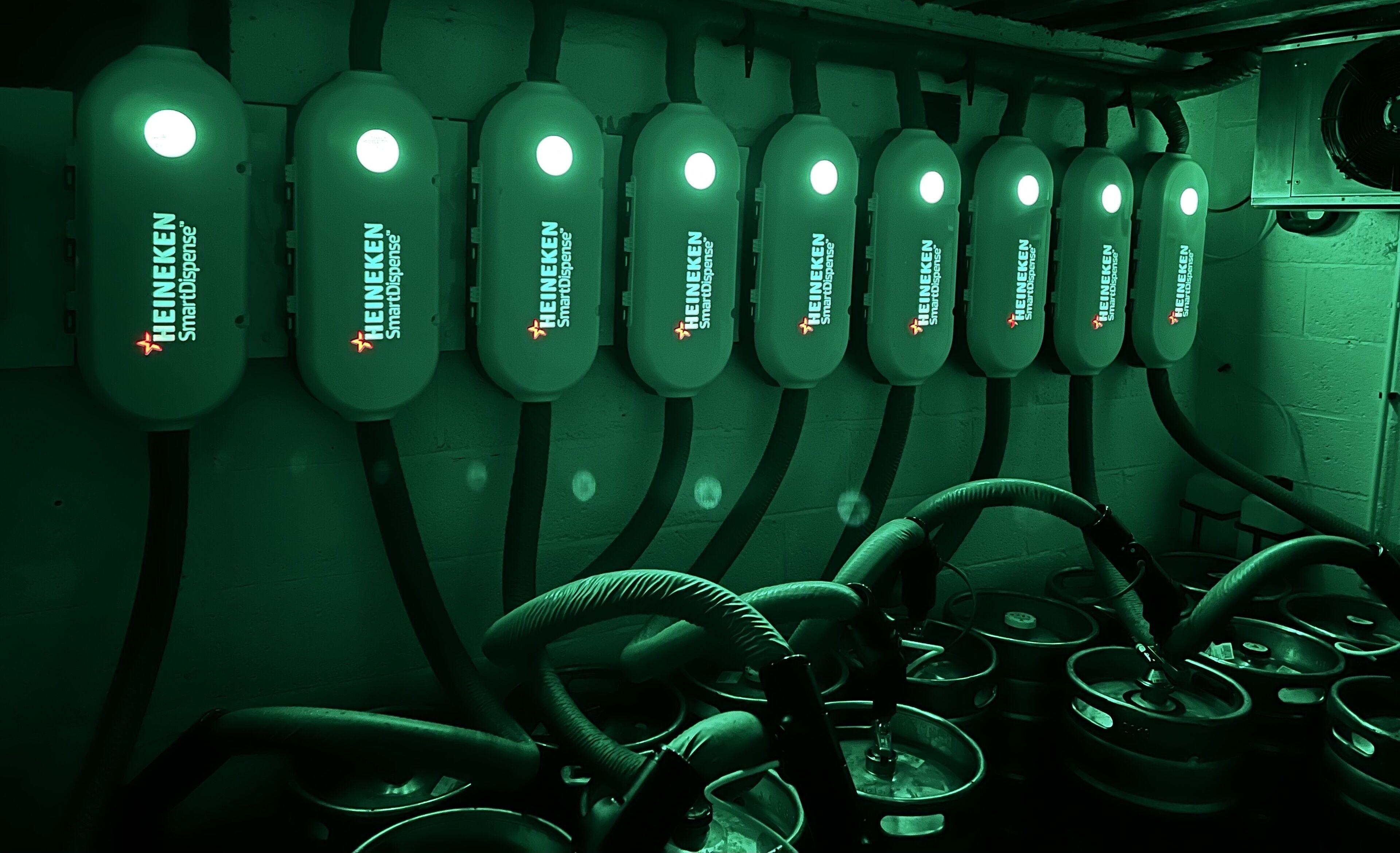 A row of HEINEKEN SmartDispense machines with hoses and kettles in a dimly lit room.