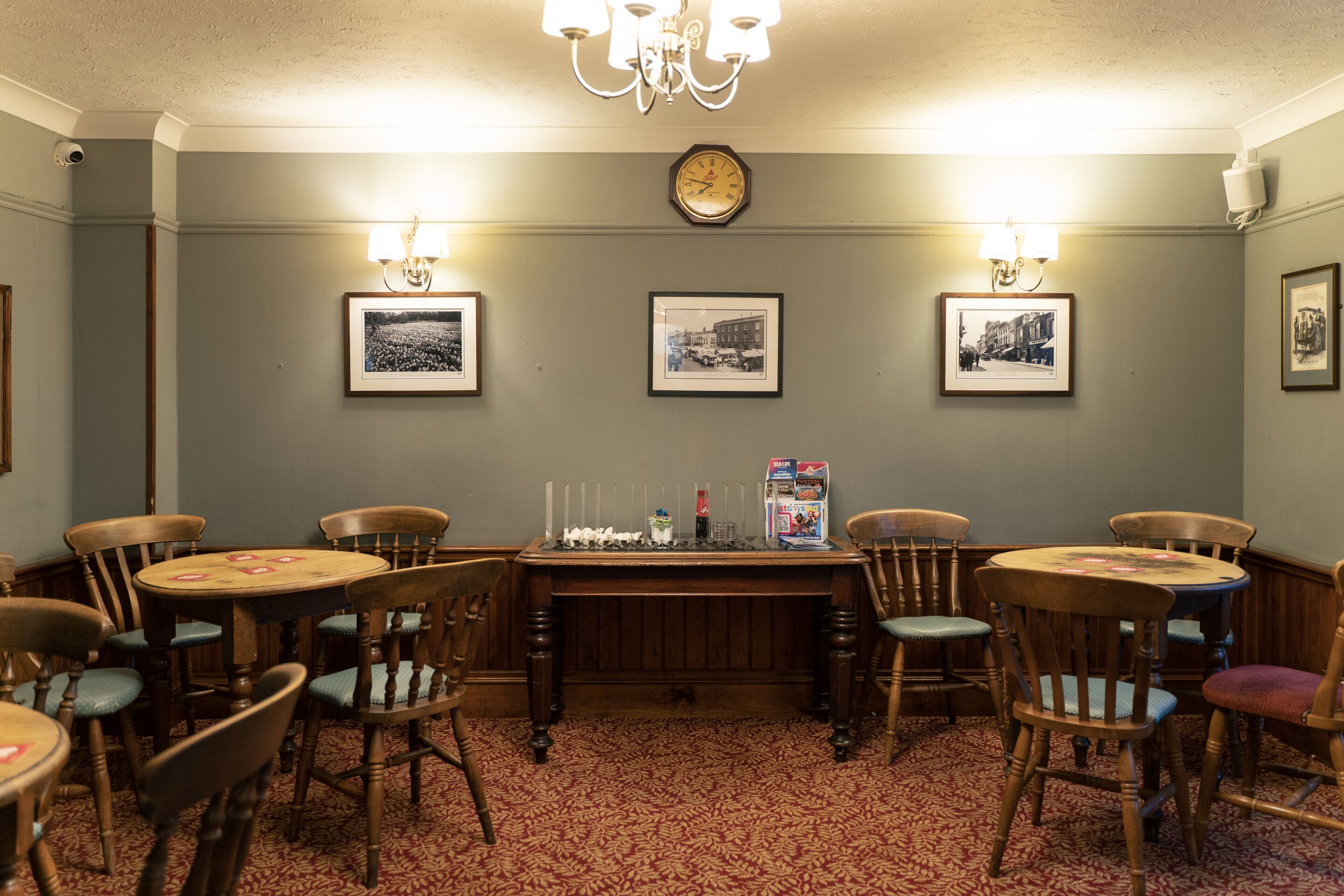 Un espace repas avec des tables et des chaises en bois, un lustre, des tableaux encadrés au mur et une horloge.