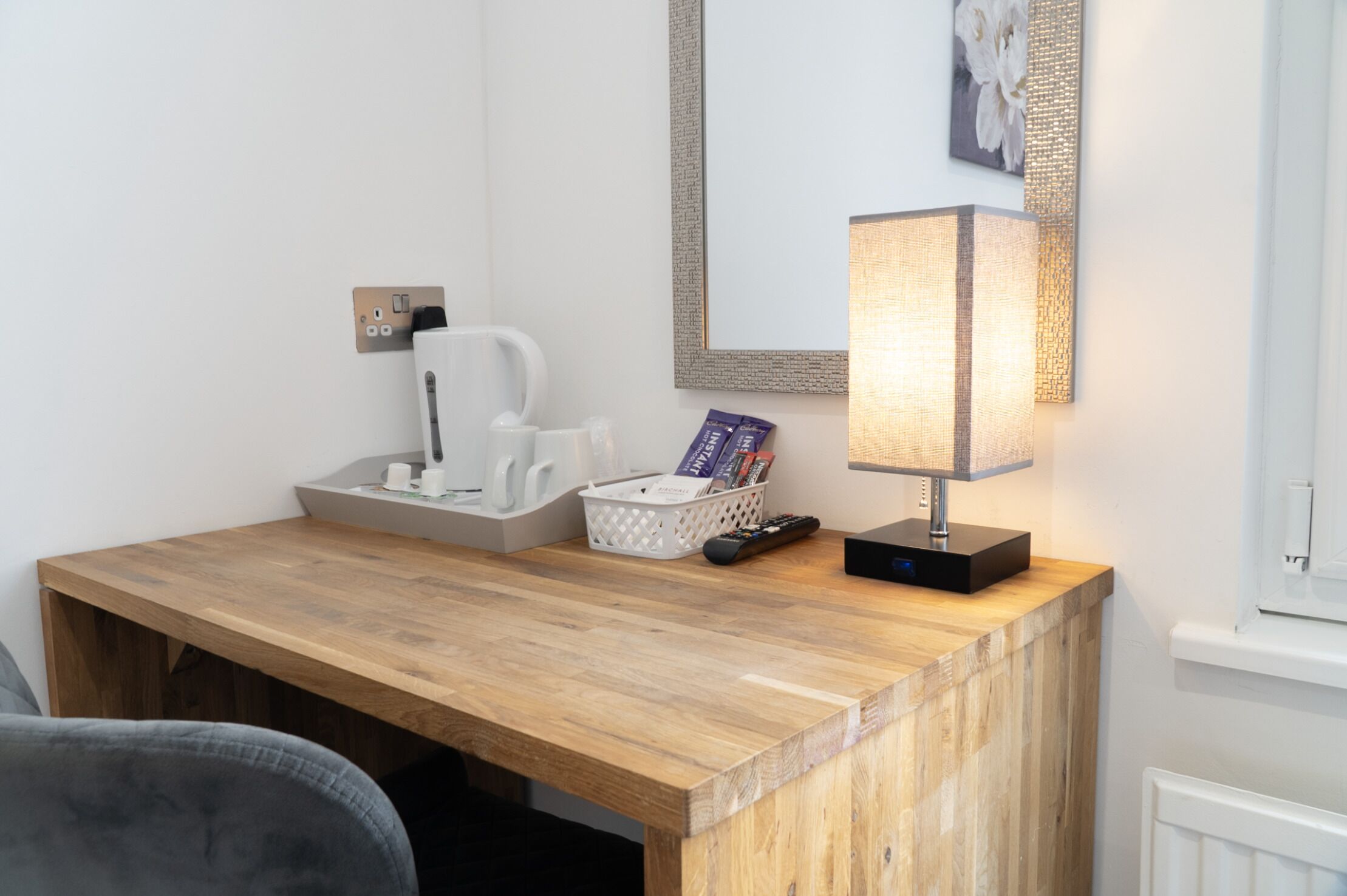A wooden desk with a lamp, a basket of books, a remote control, and a white coffee maker.