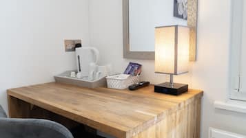 A wooden desk with a lamp, a basket of books, a remote control, and a white coffee maker.