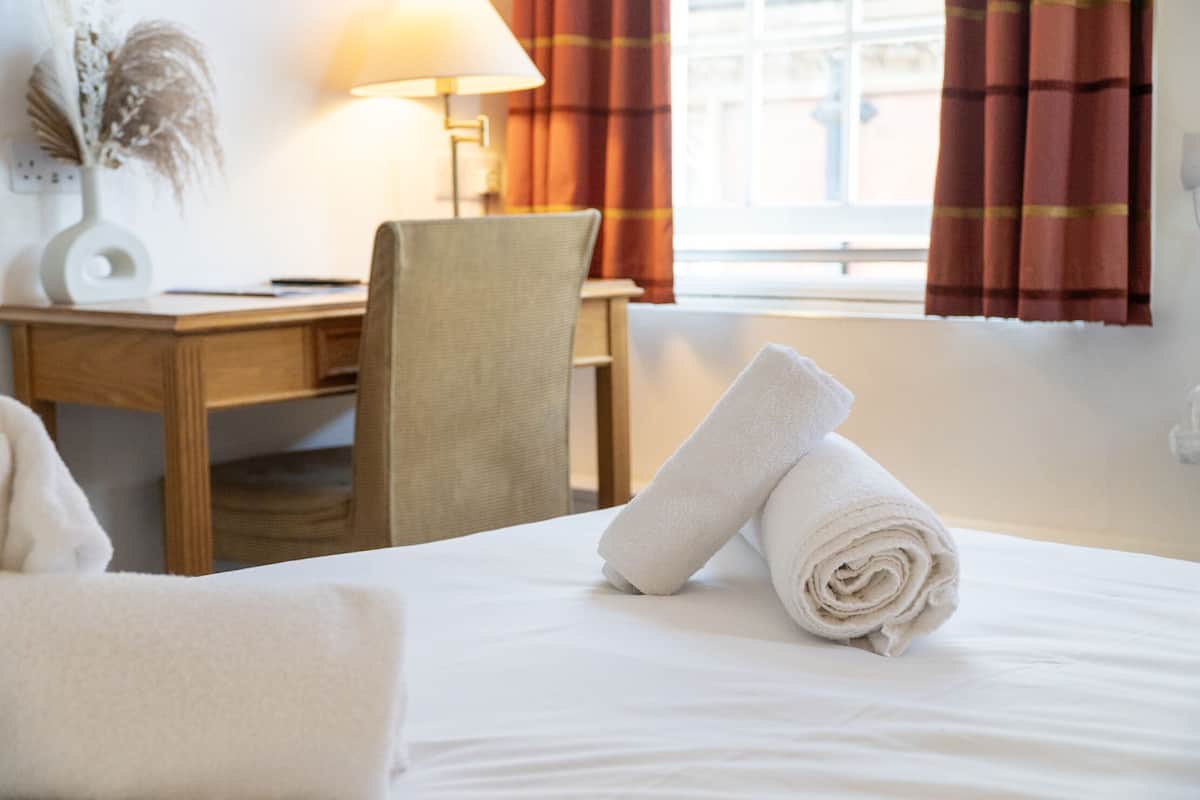 A neatly made bed with rolled white towels, a wooden desk with a lamp, and a chair in a room with a window and curtains.
