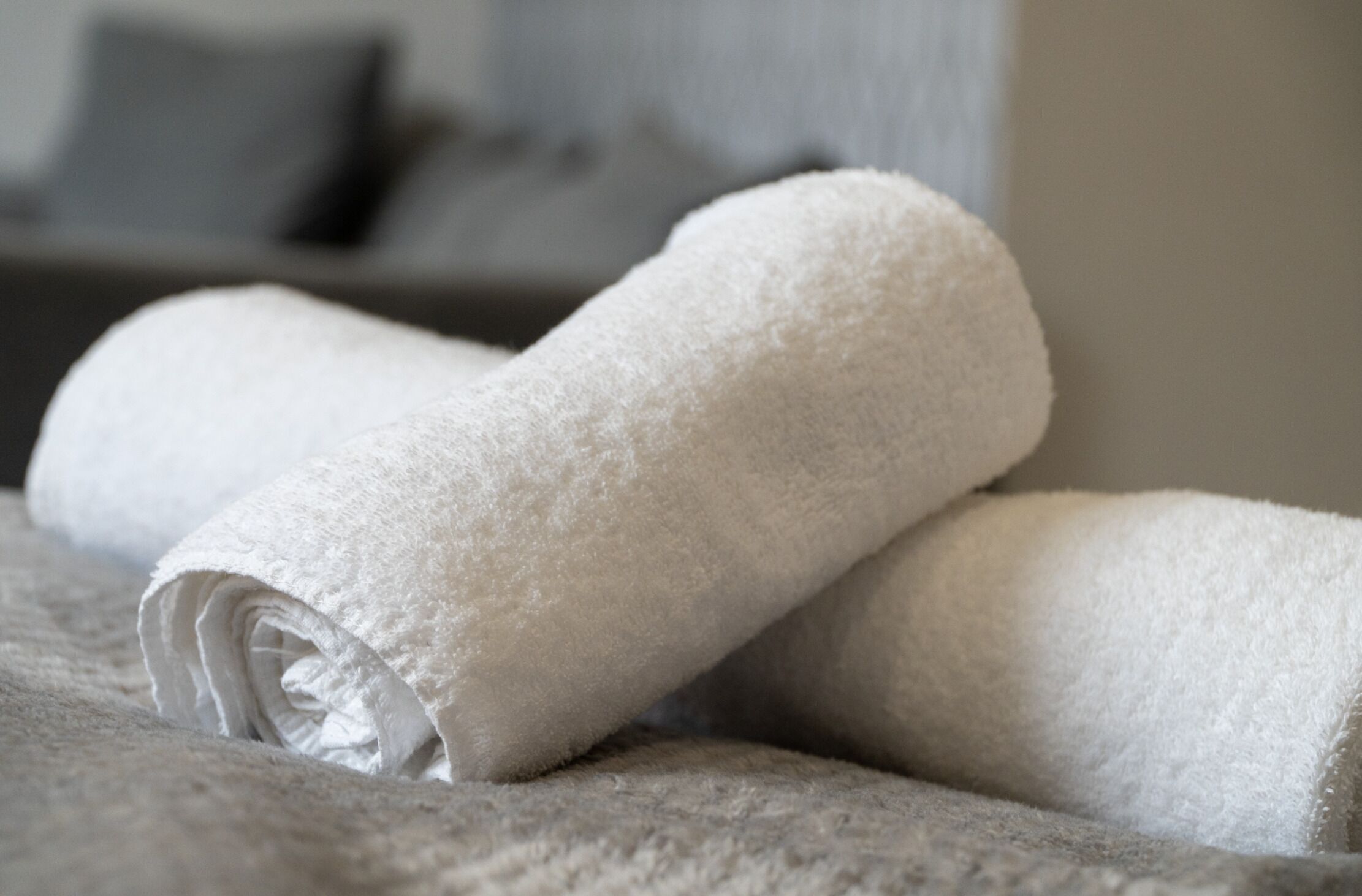 A neatly folded white towel on a textured surface, likely a bed or couch, in a room with a neutral color scheme and a blurred background.