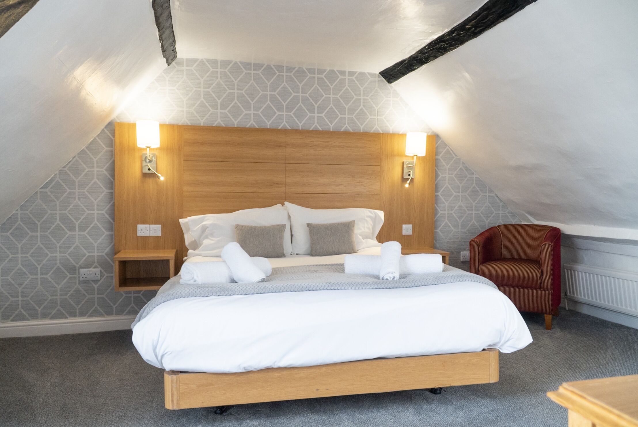 Una cama bien hecha en una habitación con techo inclinado, cabecera de madera y dos lámparas fijadas a la pared.