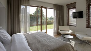 A modern hotel room with a large bed, a seating area with two chairs, a flat-screen TV, and a sliding glass door leading to a garden. - B&B Baron's House Neerijse - Leuven (Huldenberg)