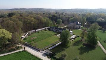 An aerial view of a large estate with expansive green lawns, a central building with a symmetrical facade, and a long, narrow structure with a glass roof.