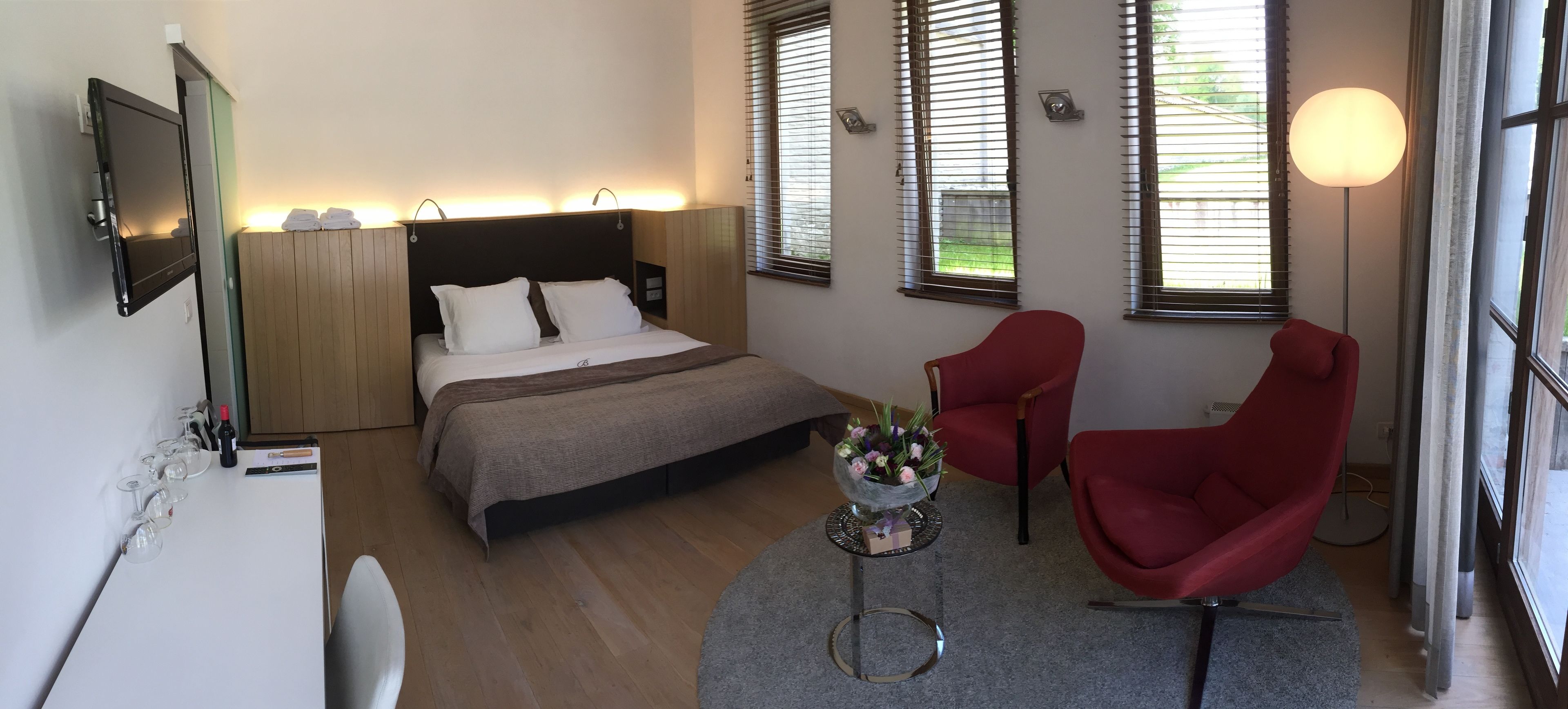 A hotel room with a bed, a red armchair, a small table with a vase of flowers, and a TV mounted on the wall.