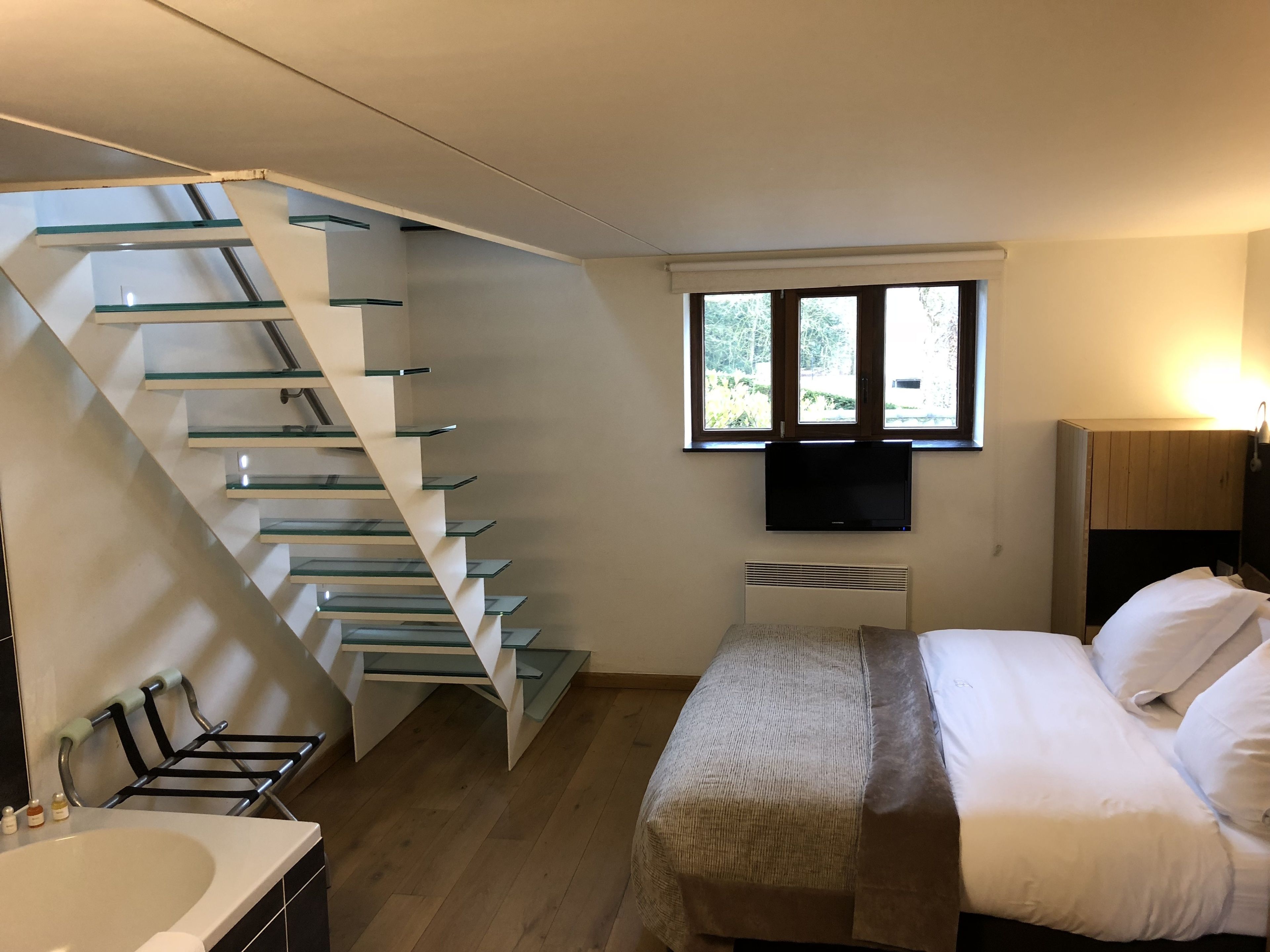Un dormitorio con una escalera moderna, una cama, un televisor y un ventanal con vistas al verdor.