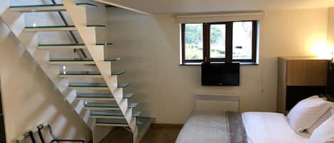 Un dormitorio con una escalera moderna, una cama, un televisor y un ventanal con vistas al verdor.