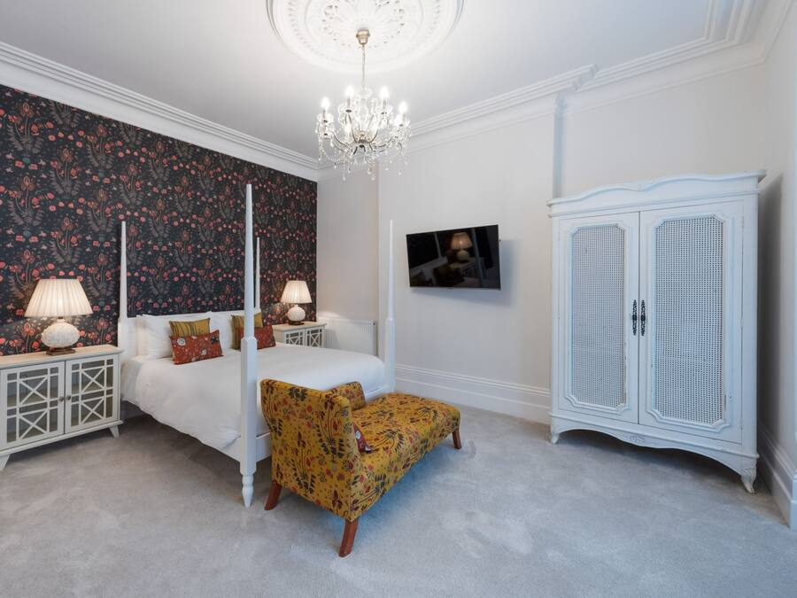 A bedroom with a four-poster bed, a nightstand with a lamp, a patterned armchair, a white wardrobe, and a floral wallpaper.