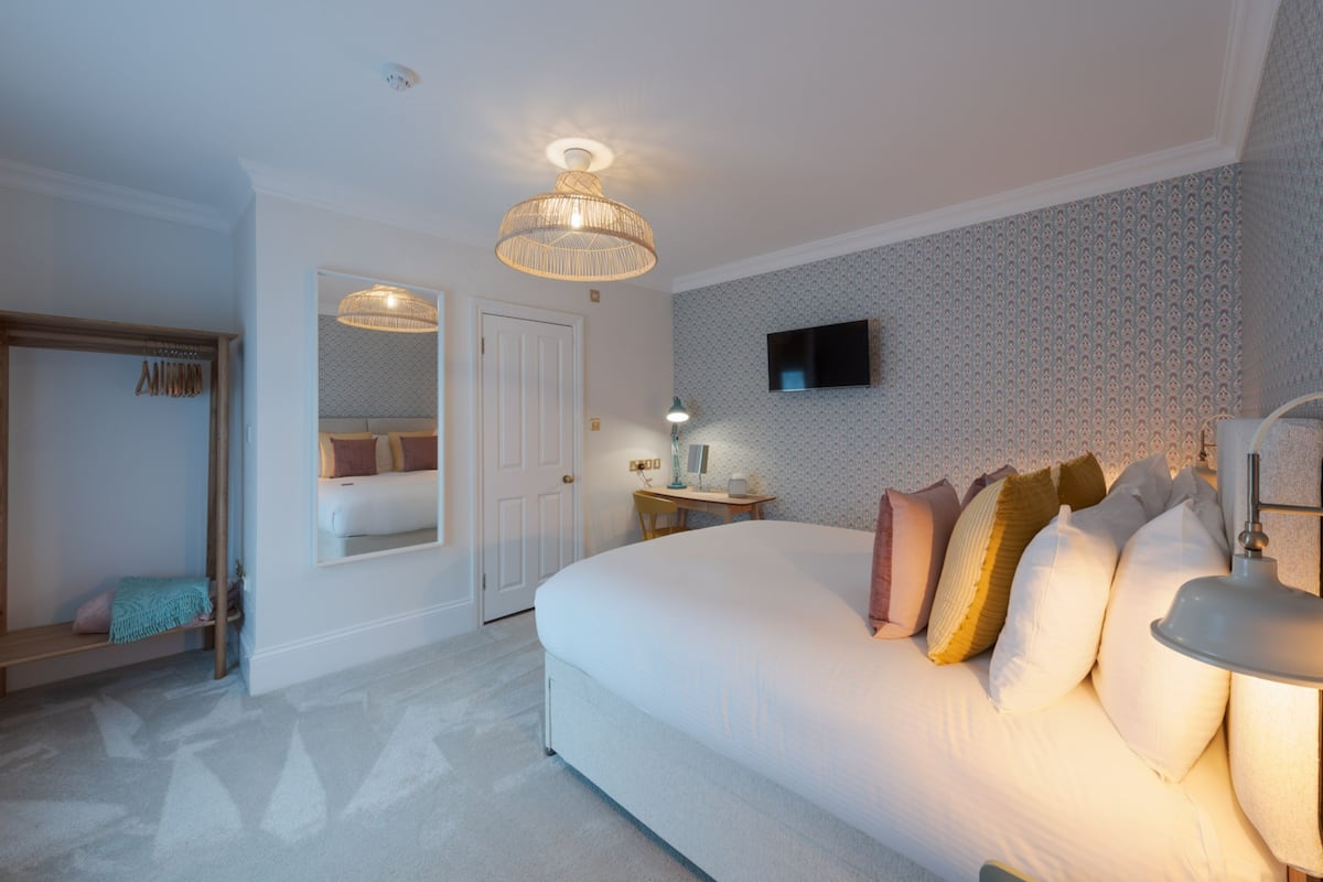 A bedroom with a large bed, a desk with a lamp, a television on the wall, and a wardrobe.