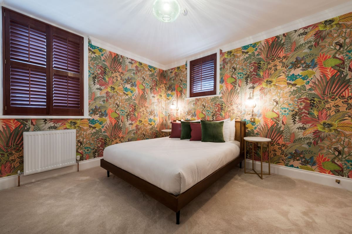 A bedroom with a large bed, floral wallpaper, a radiator, and two windows with shutters.