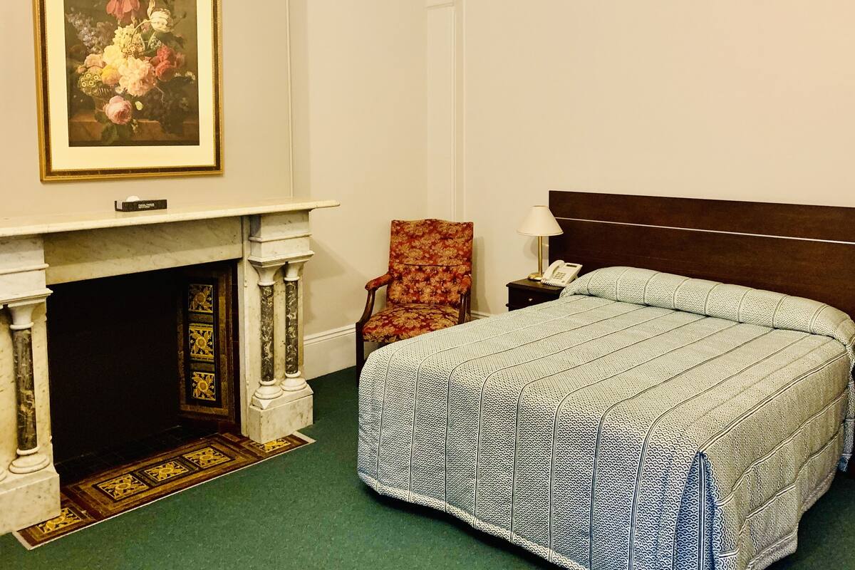 Una habitación de hotel con una cama, una silla, una chimenea y un cuadro enmarcado.
