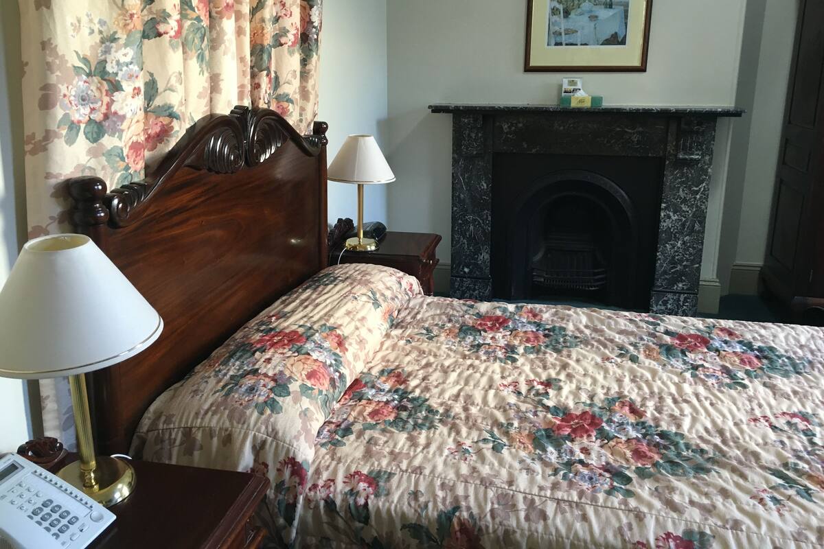 Un dormitorio con cabecera de madera, colcha estampada con motivos florales, lámpara de noche, teléfono y chimenea.