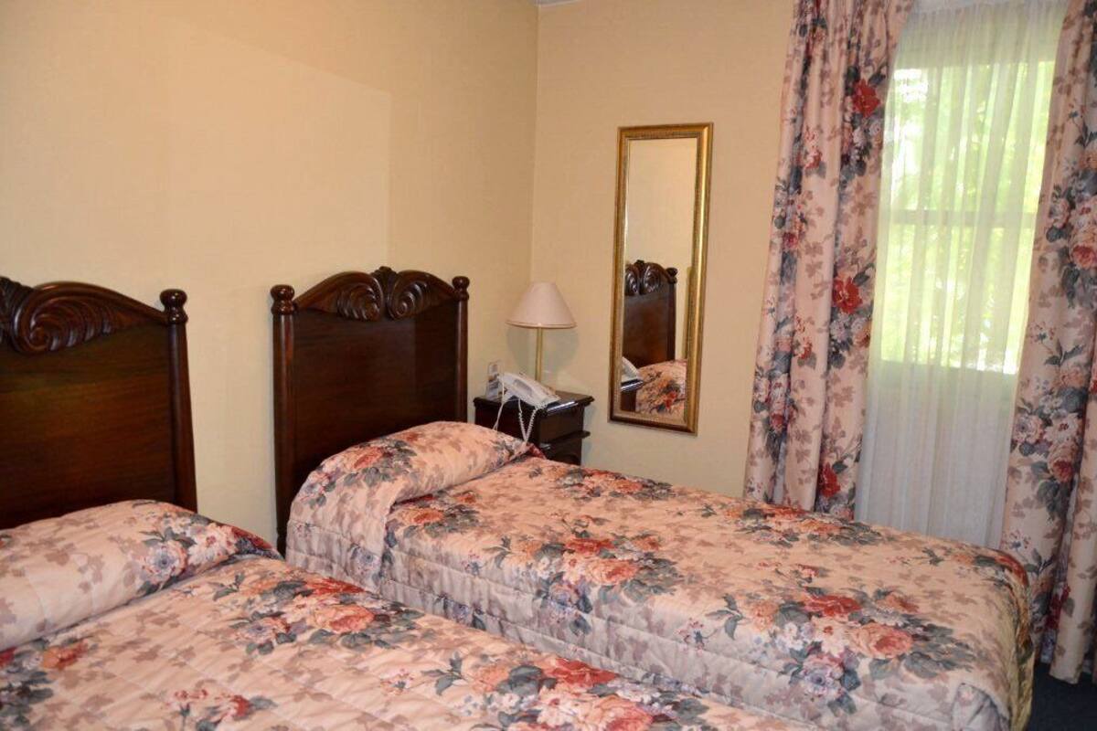 Habitación de hotel con dos camas de madera, un teléfono en la mesita de noche, un espejo y cortinas con motivos florales.