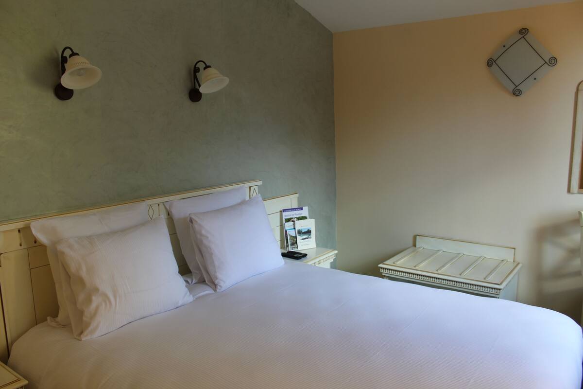 A neatly made bed with white linens, a bedside table with a lamp, and a small white nightstand.