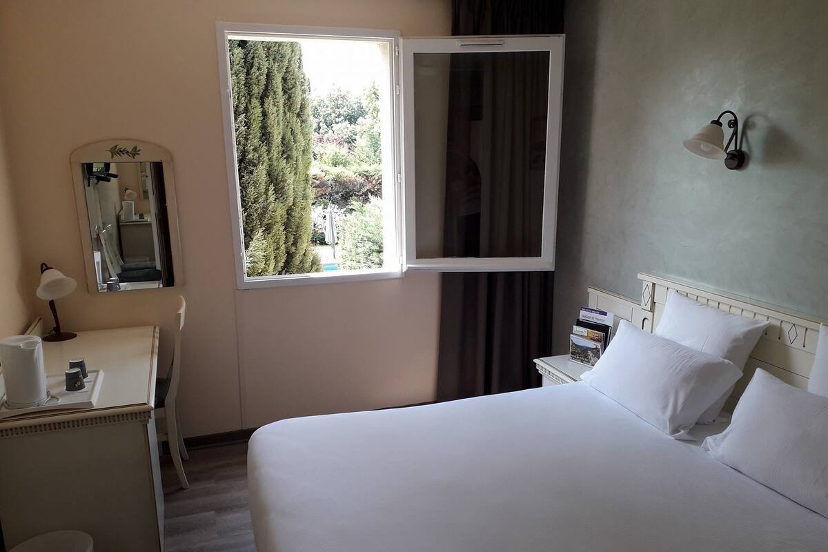 A hotel room with a bed, a desk with a lamp, a mirror, a window with curtains, and a wall-mounted light fixture.