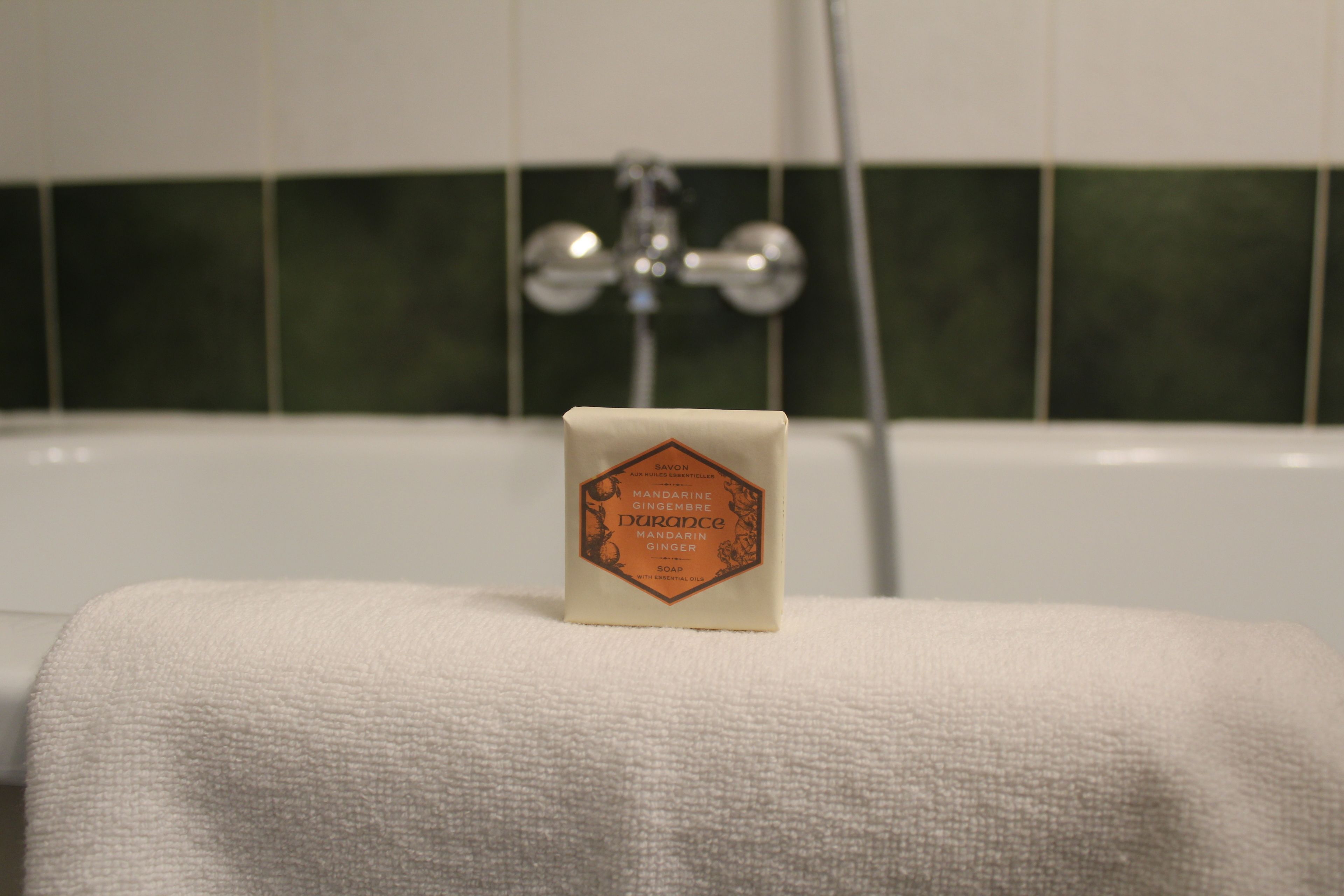 Un morceau de savon posé sur une serviette dans une salle de bain dont le mur est recouvert de carreaux verts et qui contient une baignoire blanche.