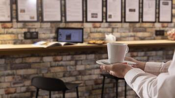 A person holding a white cup and saucer, with a brick wall and menu boards in the background.
