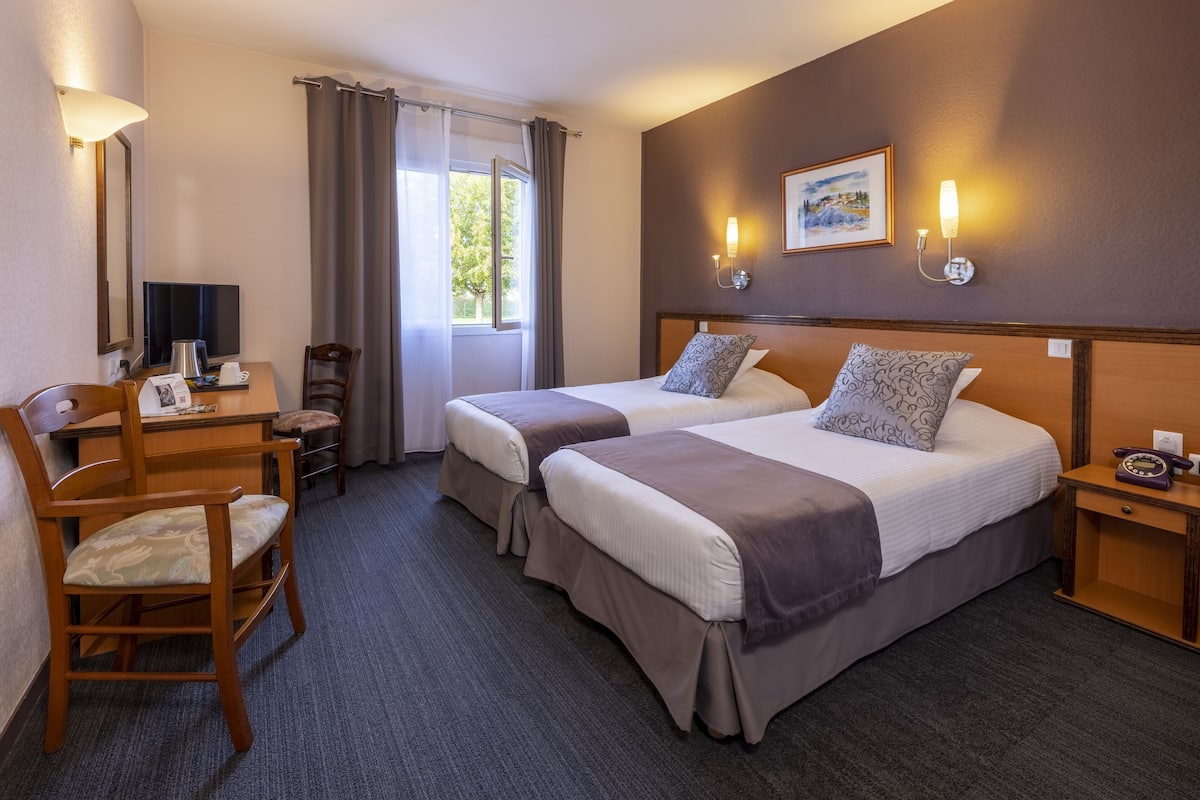 Ein Hotelzimmer mit zwei Betten, einem Schreibtisch mit Stuhl, einem Fernseher und einem Fenster mit Vorhängen.
