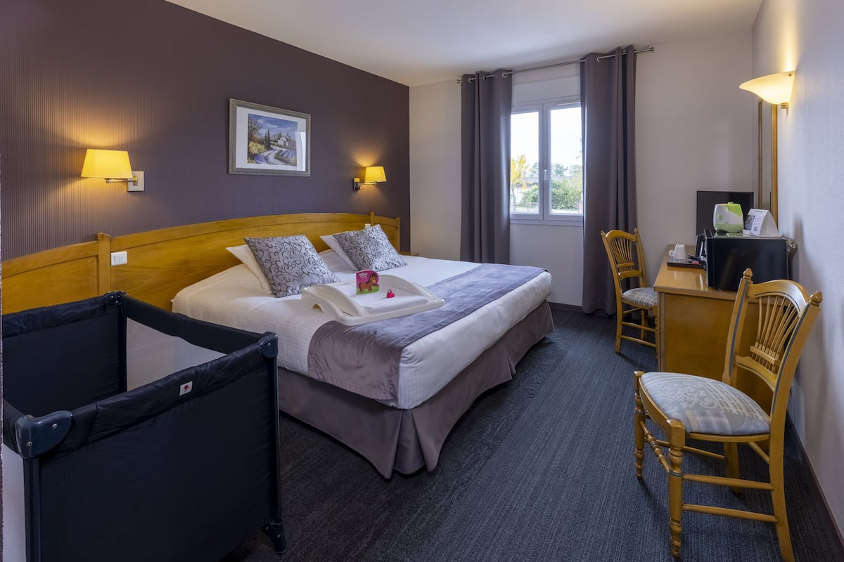Ein Hotelzimmer mit einem Bett, zwei Nachttischlampen, einem Schreibtisch mit Stuhl, einem Fenster mit Vorhängen und einem Papierkorb.