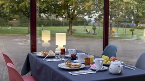 A breakfast table set with plates, cups, and a teapot, with a view of a park through large windows. - The Originals City, Hotel Paray-Le-Monial (Paray-le-Monial)