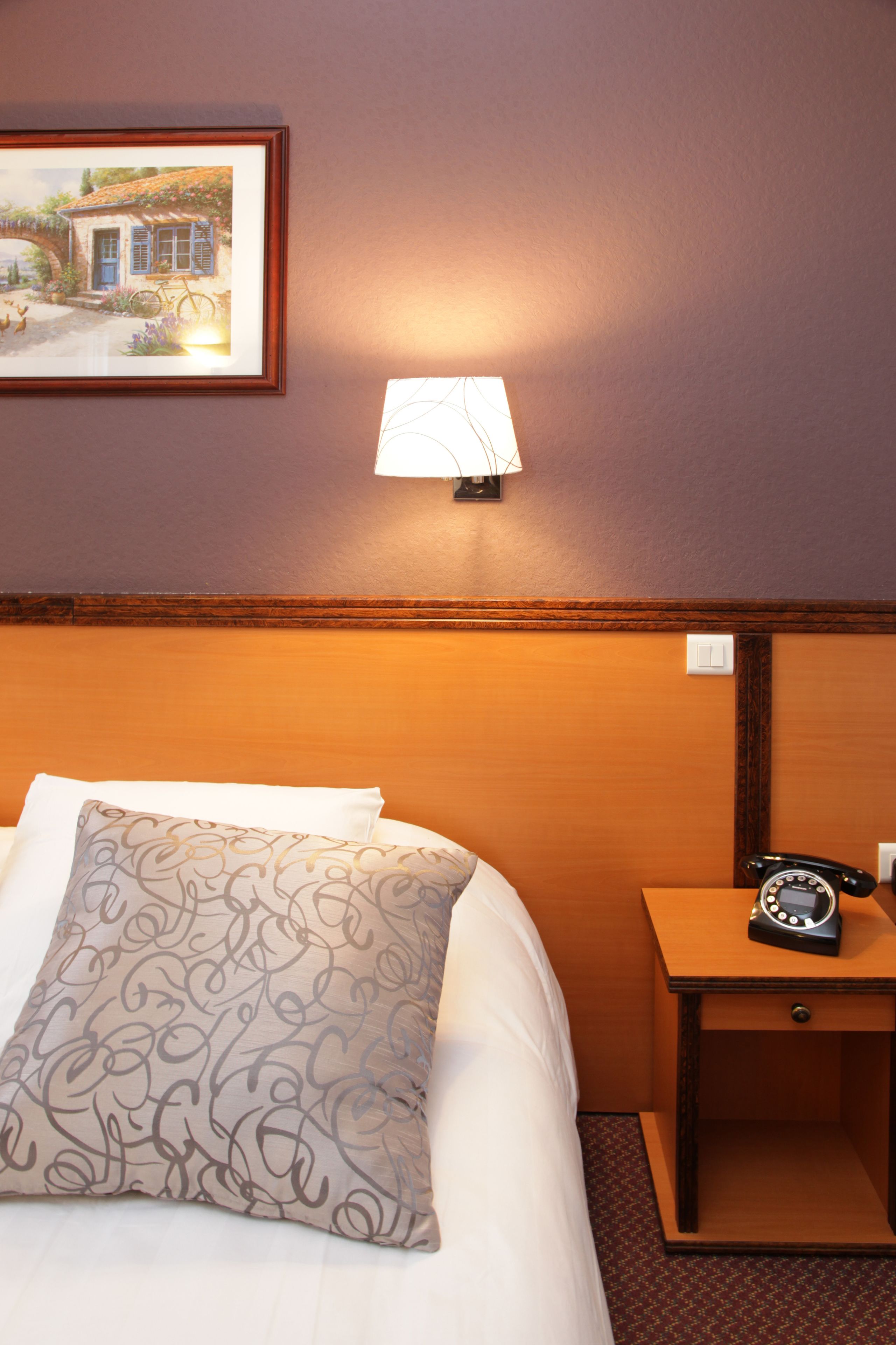 Una habitación de hotel con cama, mesita de noche con teléfono, lámpara de pared e imagen en un marco colgado en la pared.