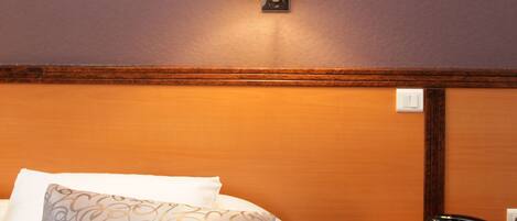 Una habitación de hotel con cama, mesita de noche con teléfono, lámpara de pared e imagen en un marco colgado en la pared.