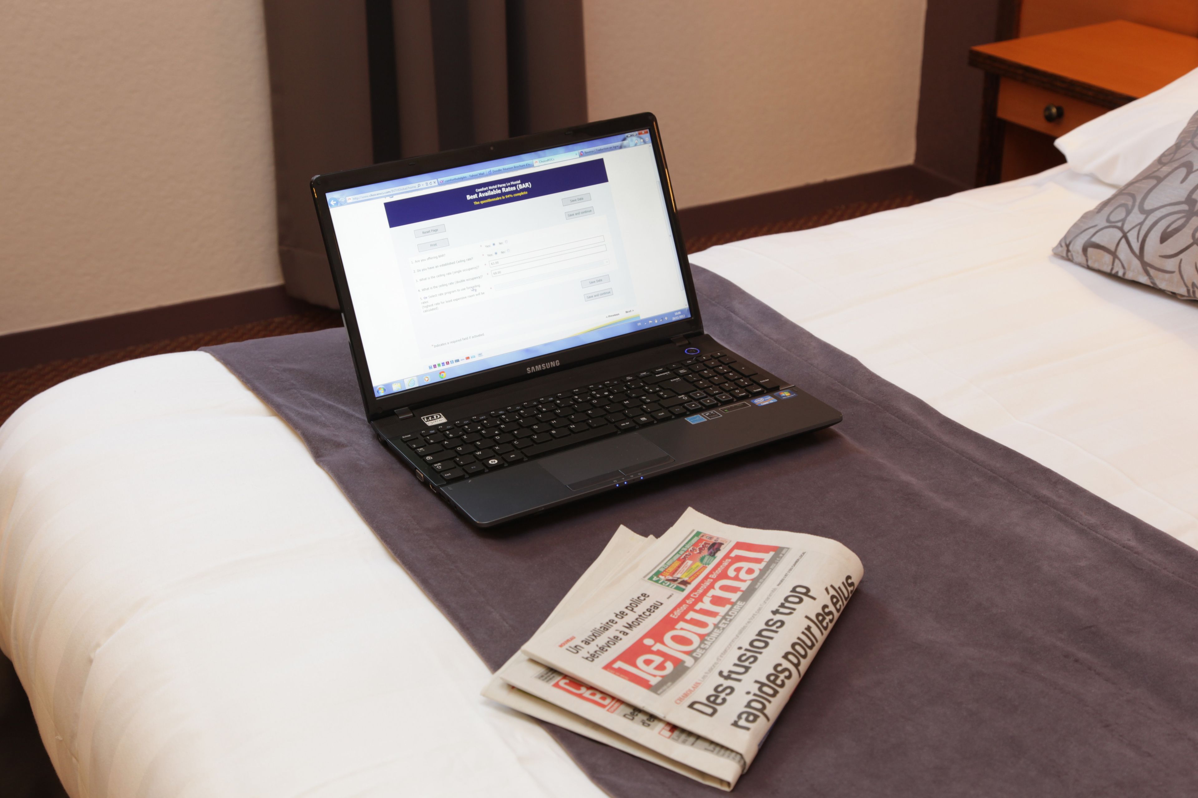 Un ordinateur portable sur un lit, avec un journal et un oreiller.