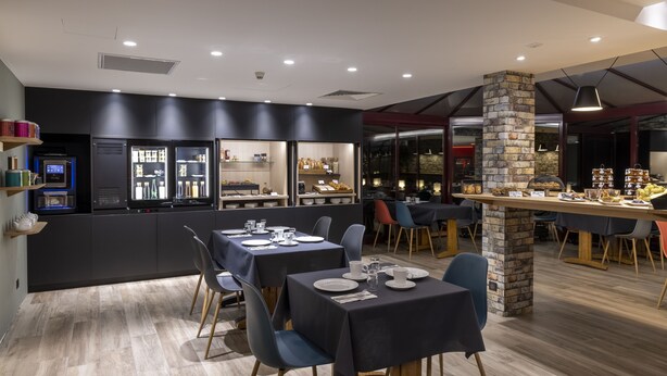 A modern restaurant with dark interiors, wooden flooring, and a stone pillar. Tables are set with plates and glasses, and there are shelves with food items. - The Originals City, Hotel Paray-Le-Monial (Paray-le-Monial)