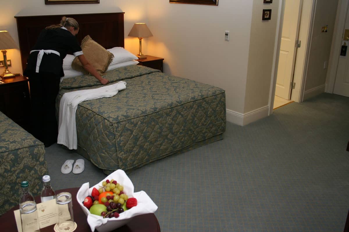A hotel room with two beds, a nightstand with a lamp, a fruit basket, and a bottle of wine on the table.