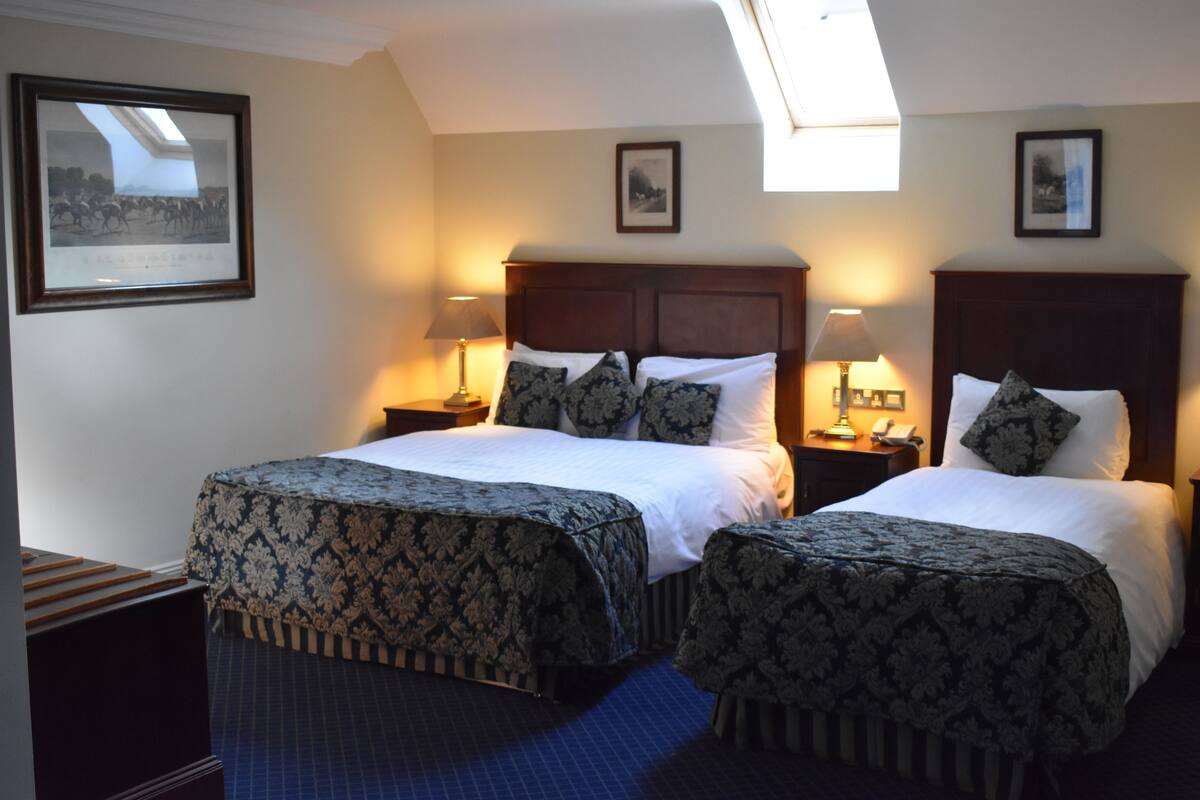 A hotel room with two beds, a skylight, framed pictures on the walls, and a small table with a lamp.