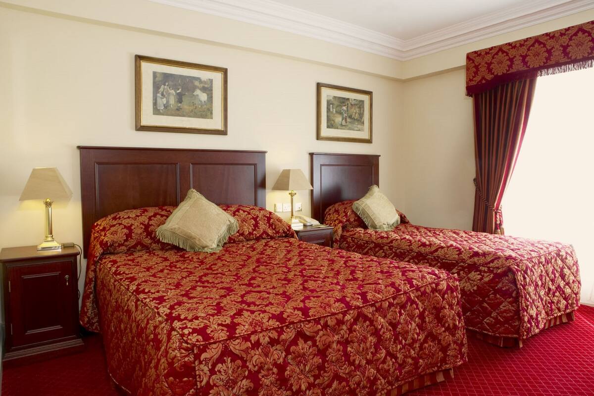 A hotel room with two beds, each with a wooden headboard, a red patterned bedspread, and a beige pillow. There are two framed pictures on the wall, a lamp on the nightstand, and a red carpeted floor.