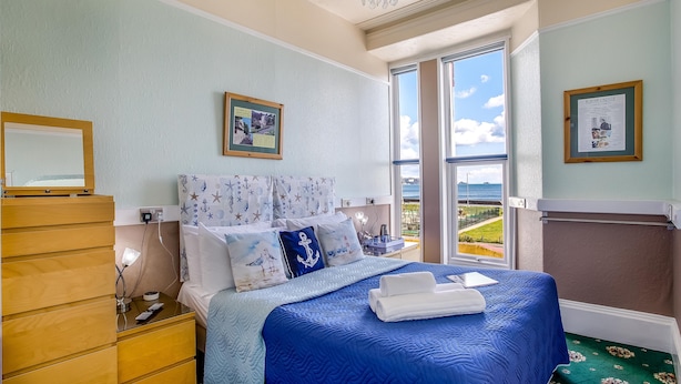 A bedroom with a large bed, a wooden dresser, a nightstand, a chandelier, and a view of the sea. - The Commodore (Paignton)