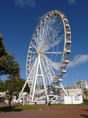 A large Ferris wheel with cabins, surrounded by a park area with trees and a historic building in the background.