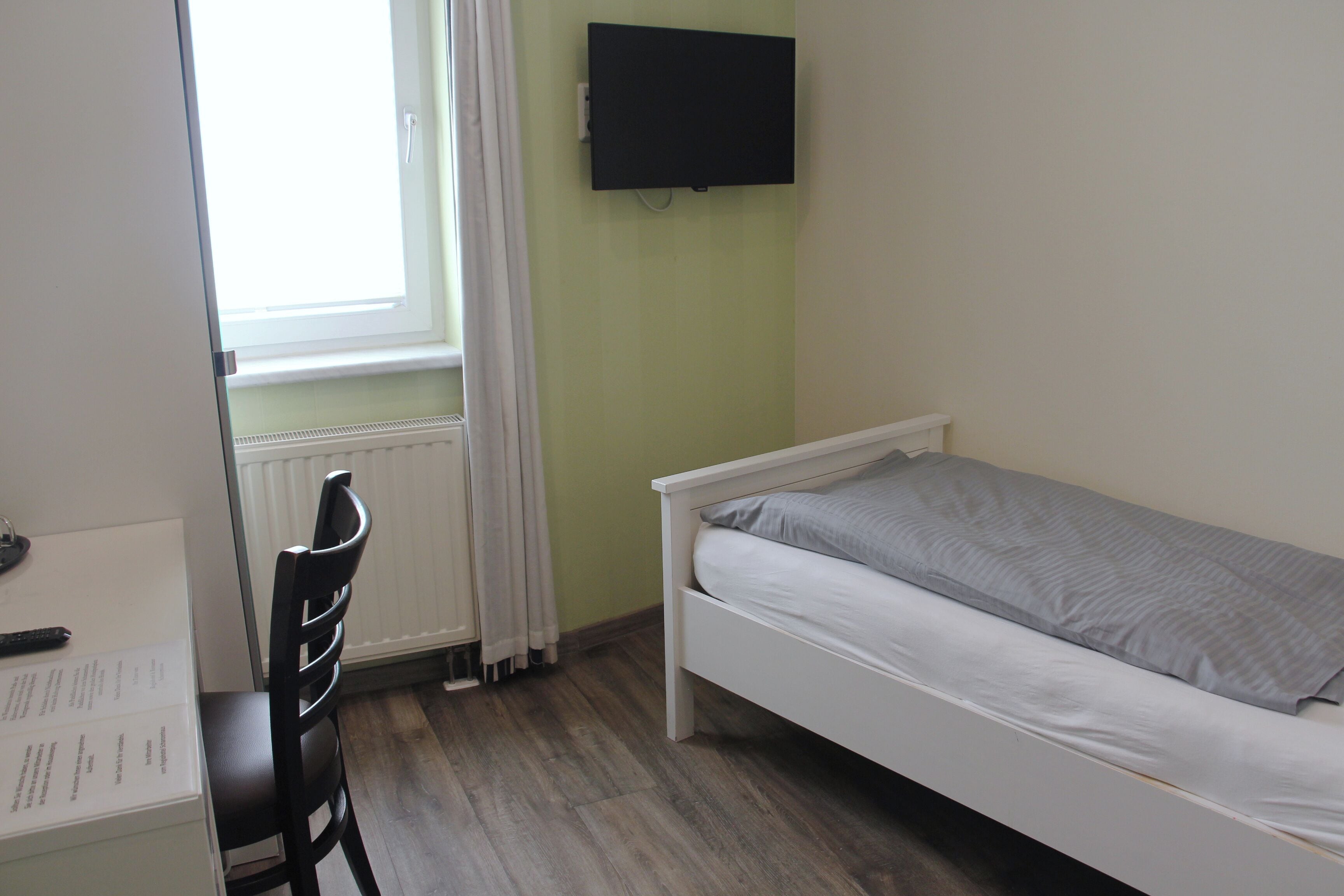 A small, single-bed room with a white bed, a desk with a chair, a flat-screen TV mounted on the wall, and a window with curtains.