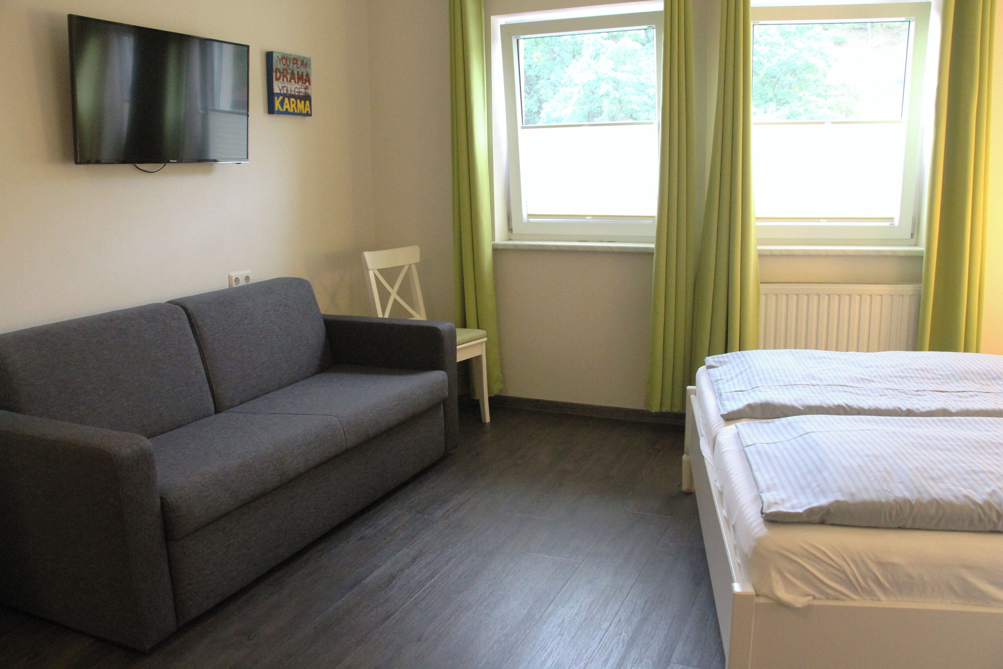 Een kamer met een grijze bank, een klein wit tafeltje, een bed met wit beddengoed, een aan de muur bevestigde televisie en een raam met groene gordijnen.