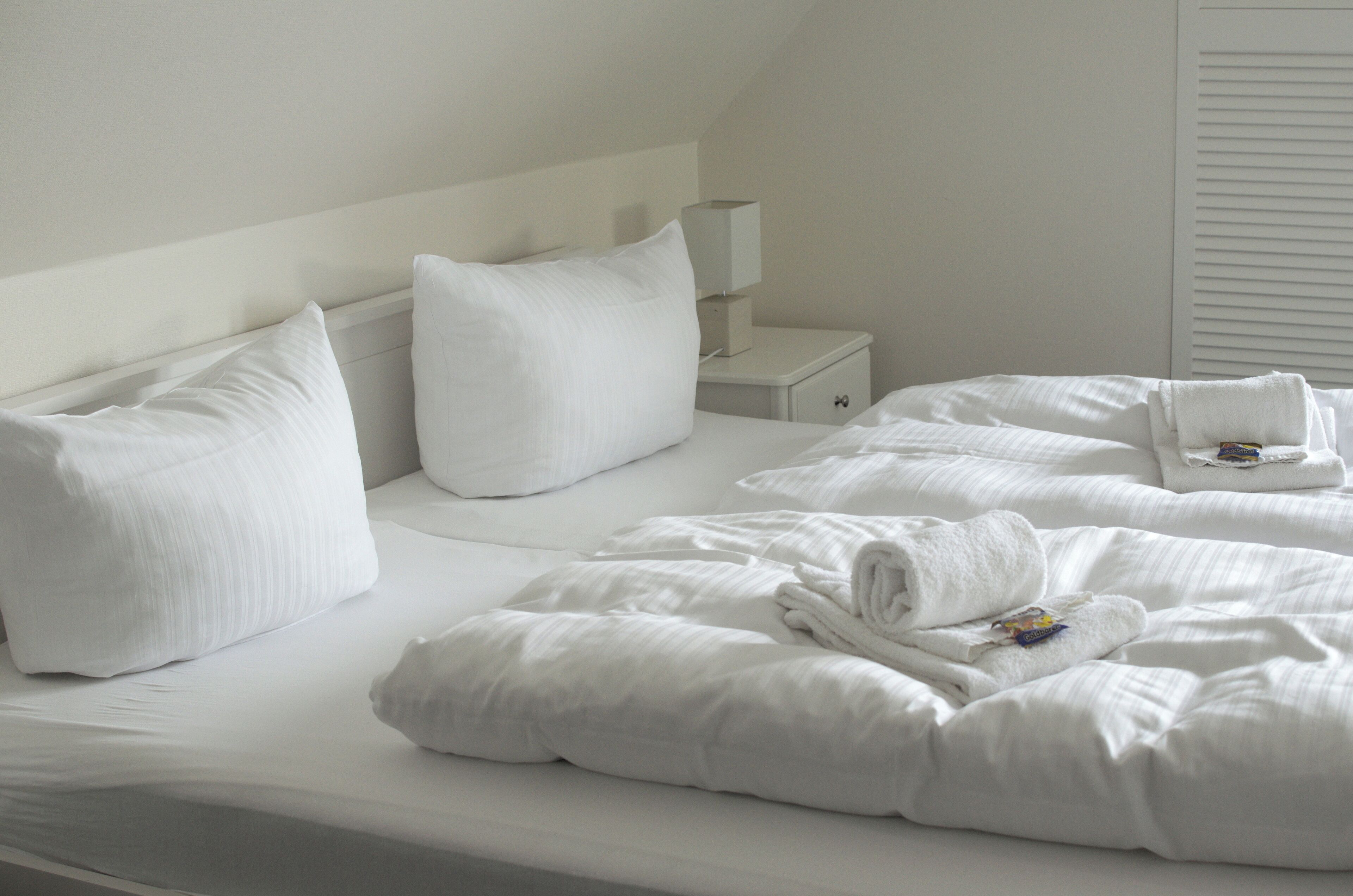 Una cama bien hecha con sábanas y almohadas blancas, una mesita de noche con una lámpara, y una ventana con persianas.